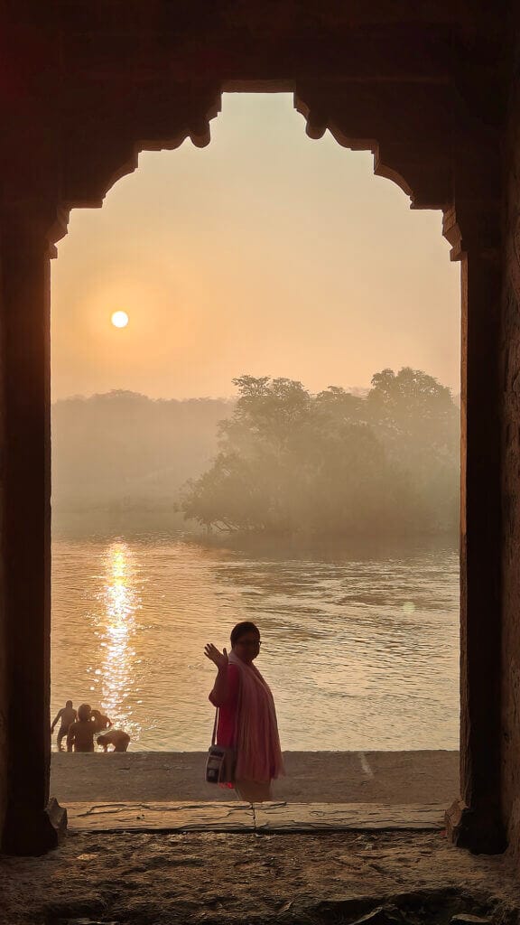 A person in a pink outfit waves while standing in an arched doorway, with a river, trees, and the sun reflecting on the water in the misty background at sunrise or sunset.