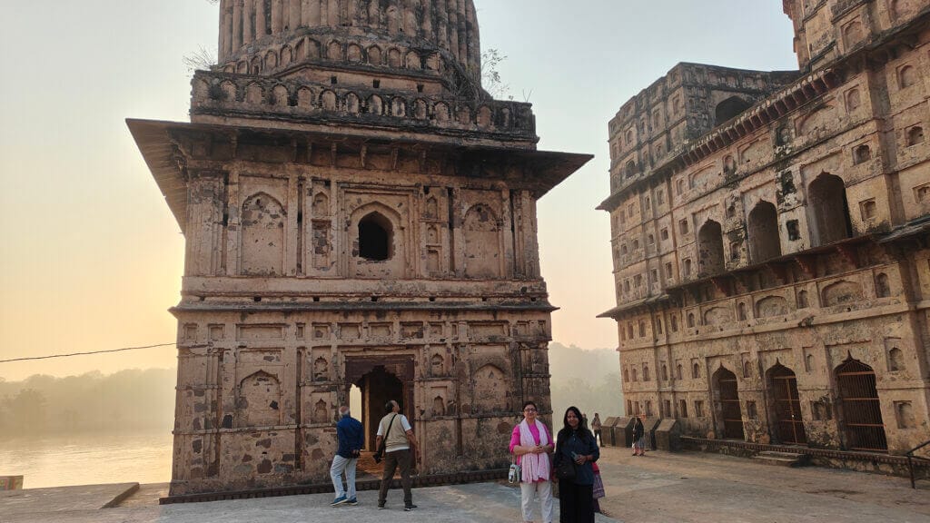 Four people stand near ancient, weathered stone buildings by a river at sunrise or sunset, with two women in the foreground and two people looking at the structure in the background. The sky is hazy and golden.