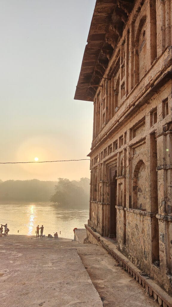 A sunrise scene by a river, with an old stone building on the right, silhouettes of people near the water, and trees in the background reflecting the sun’s light.