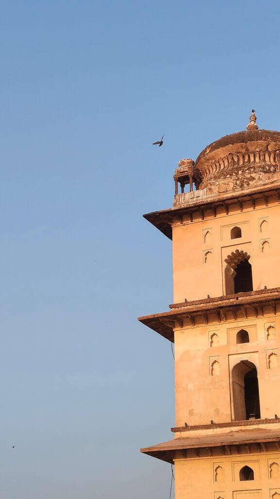 A portion of an old, ornate sandstone building with arched windows and detailed carvings against a clear blue sky, with a bird flying near the upper dome.