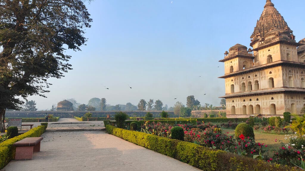 A historic sandstone building with ornate towers stands in a manicured garden with flowers, shrubs, and pathways. Trees and more historic structures are visible in the background under a clear blue sky.