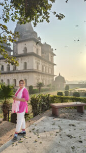 A woman in a pink kurta and white pants stands in front of a historic stone building with domed towers, surrounded by greenery. Birds fly in the sky, and sunlight creates a soft glow in the scene.