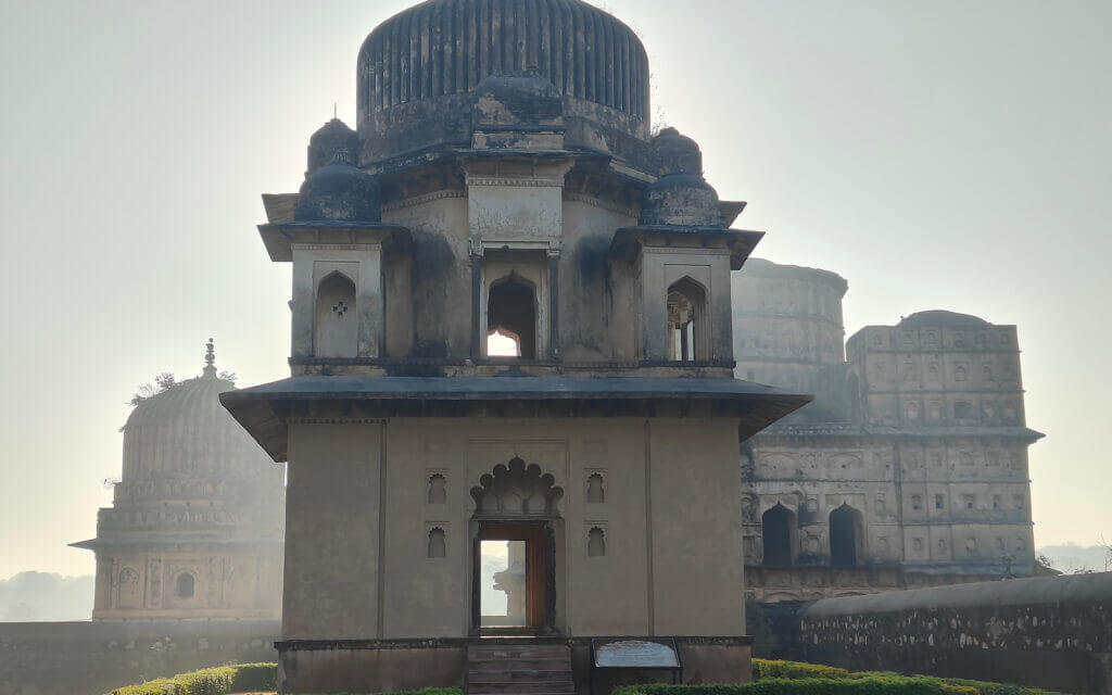 Historical stone monument with arched windows and domed roof, set against a bright sky; two other domed structures are visible in the background, partially obscured by mist.