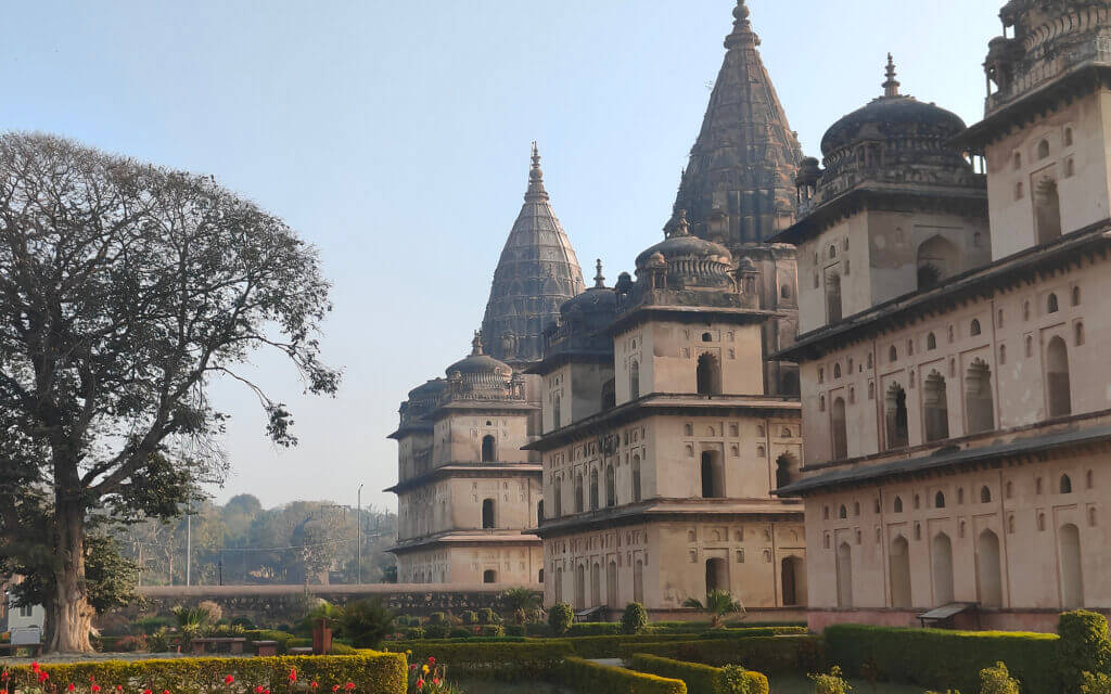 Historic stone temples with tall spires and intricate carvings, surrounded by manicured gardens, a large tree on the left, and a clear blue sky in the background.