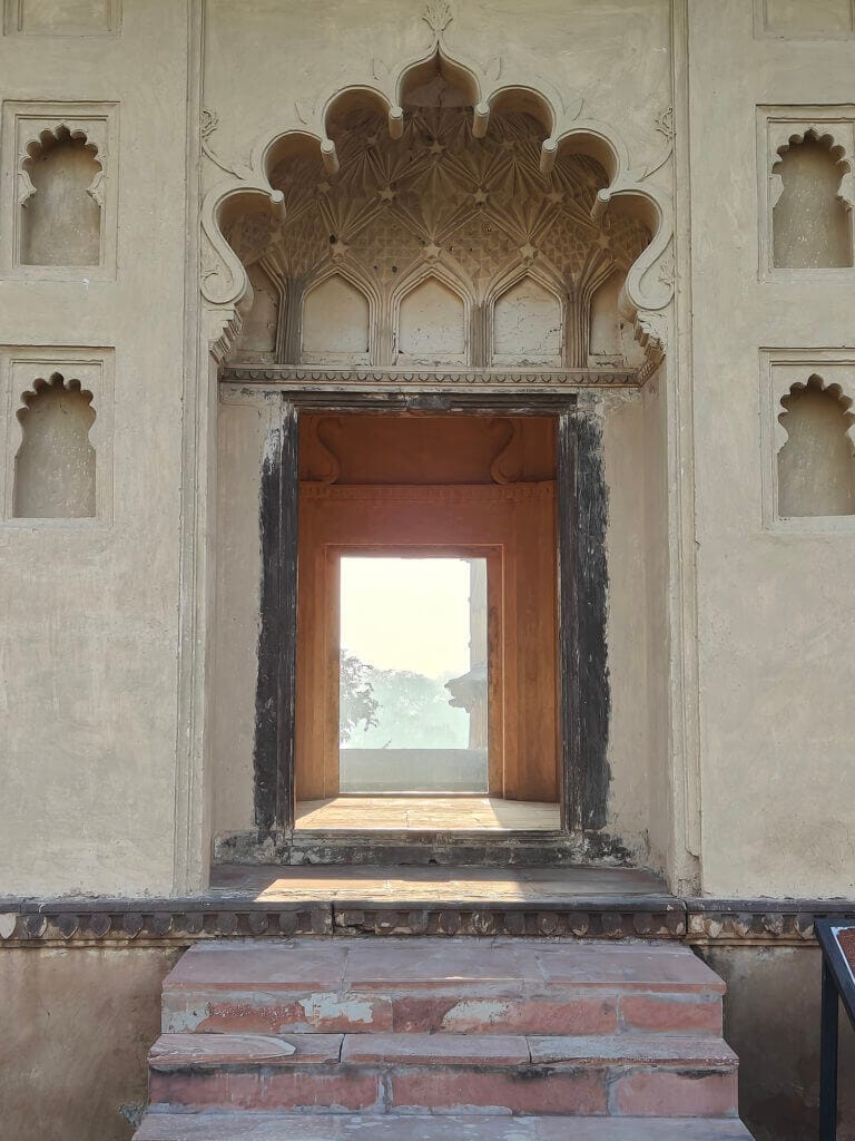 An arched doorway with intricate carvings, surrounded by small decorative niches, opens to a view of another arched frame and a sunlit landscape beyond; brick steps lead up to the entrance.