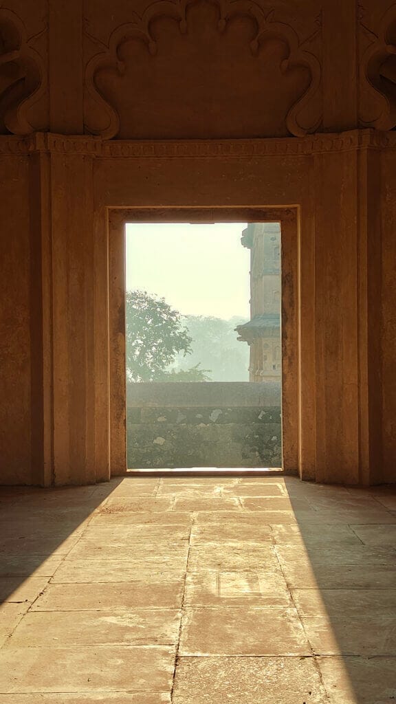 Sunlight streams through an ornate window and a doorway of an ancient, arched sandstone structure, casting warm light on the textured walls. A hazy tree and distant architecture are visible through the doorway.