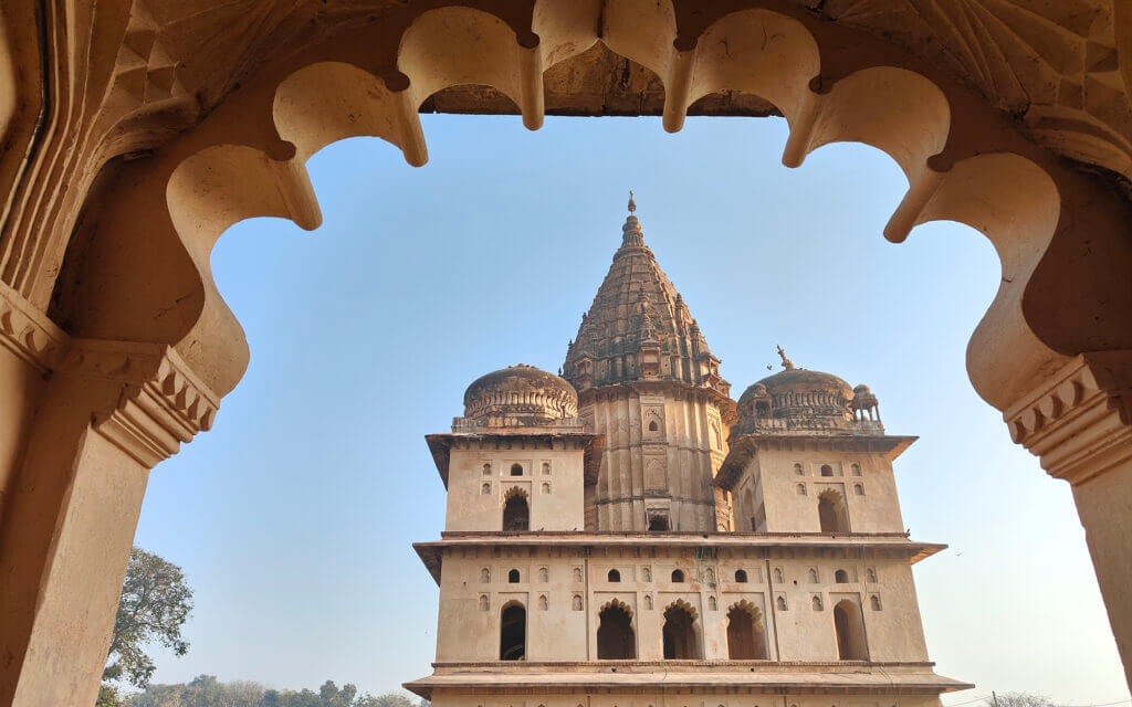 A historic temple with ornate domes and a tall central spire is framed by an arched window with intricate carvings, against a clear blue sky.