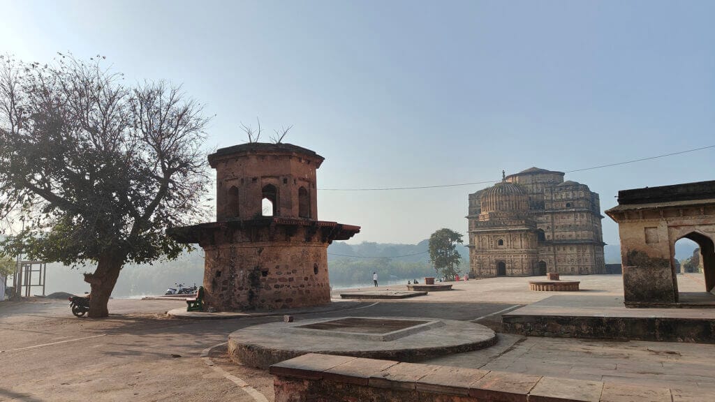 A historic stone structure and a round tower stand in an open courtyard, with a leafless tree nearby and a large building in the background under a clear blue sky.