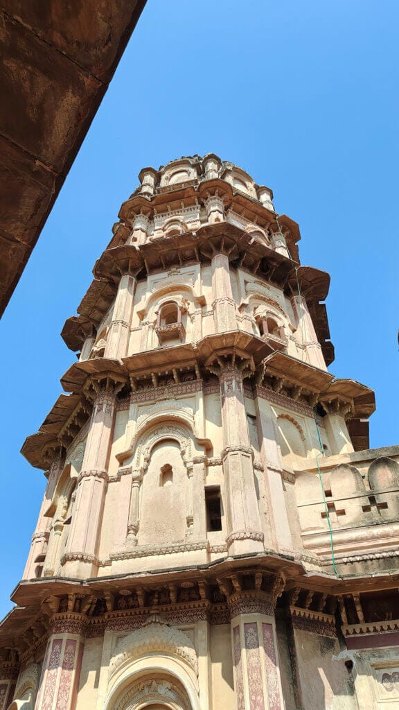 A tall, intricately carved stone tower with multiple levels and arched windows, seen from below against a clear blue sky. The architecture features ornate details and faded pinkish-beige tones.