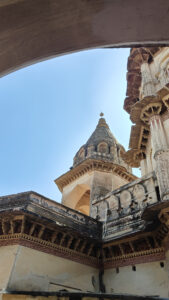 A low-angle view of an old temple with ornate domes and intricate architectural details, set against a clear blue sky, partially framed by an archway.
