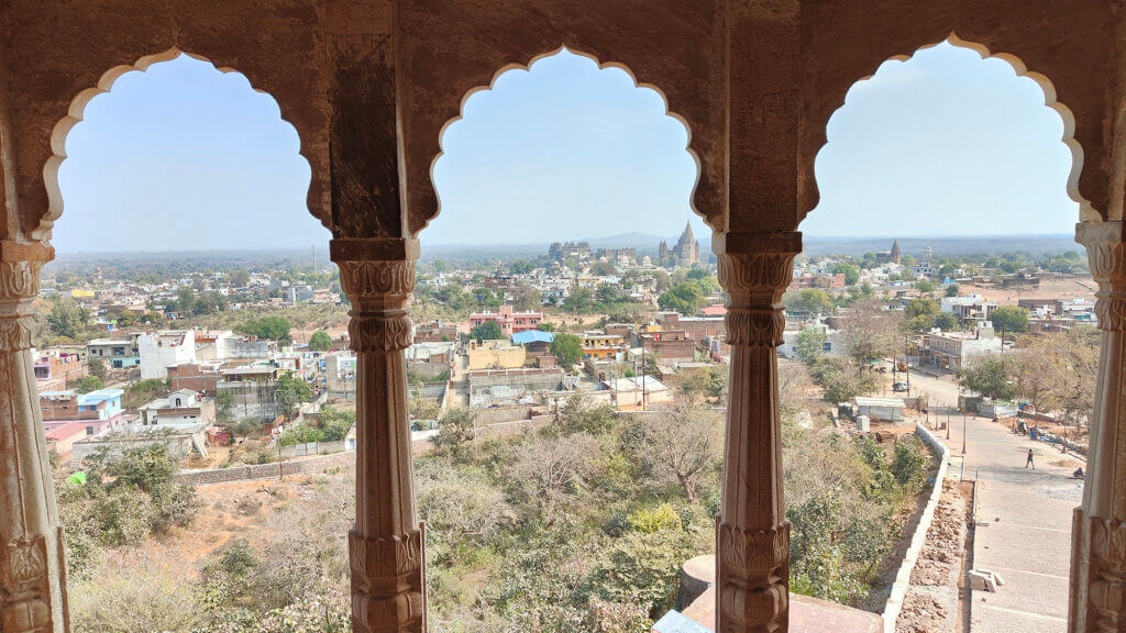 View of a town with historic buildings and temples seen through three ornate stone arches, overlooking a mix of greenery and rooftops under a clear sky.