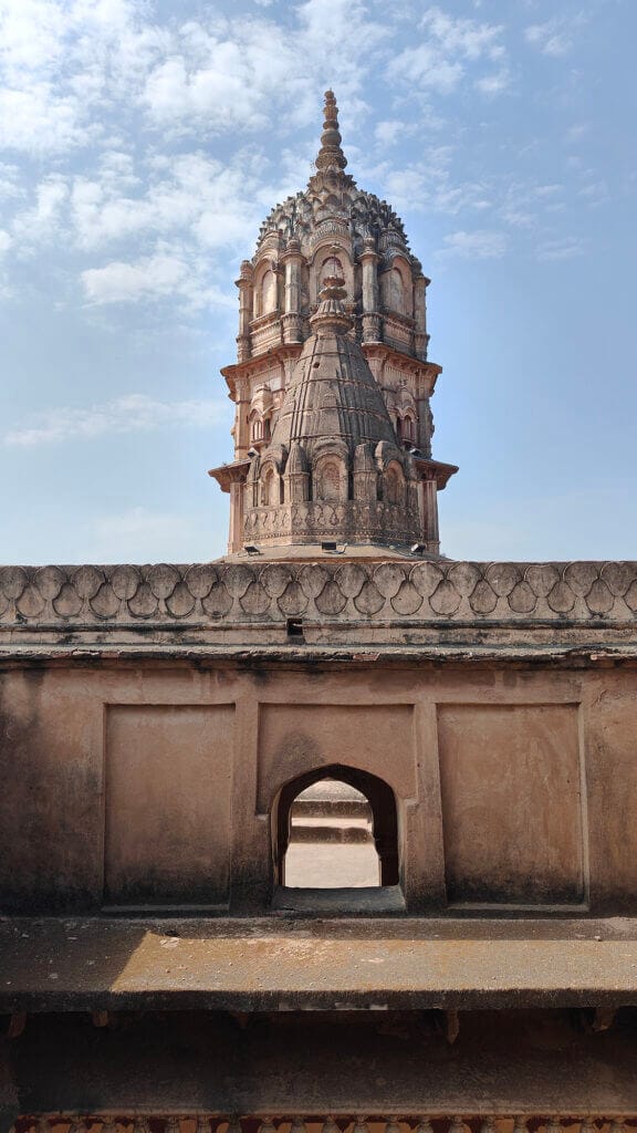 A historic stone temple with an ornate, tiered tower rises behind an old, weathered wall featuring a small archway, under a partly cloudy blue sky.