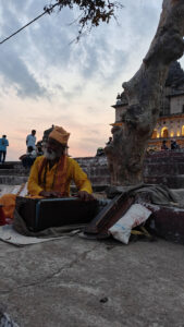 A bearded man in traditional attire plays a harmonium near a tree at sunset, with a temple and people in the background. The scene is peaceful and evocative, capturing a cultural moment outdoors.