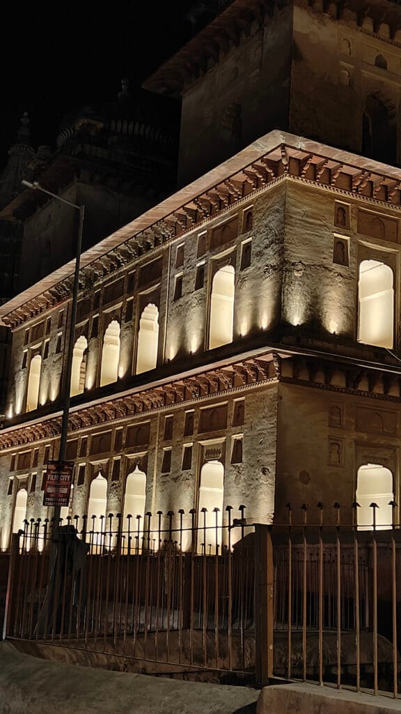 A historic building with arched windows and intricate carvings is illuminated at night by warm lights, highlighting its architectural details. A metal fence runs along the front of the structure.